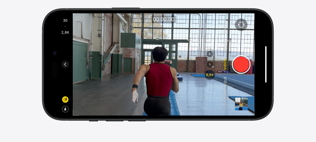 Image d’un iPhone 15 Pro qui filme en mode Action un gymnaste courant dans une grande salle lumineuse.