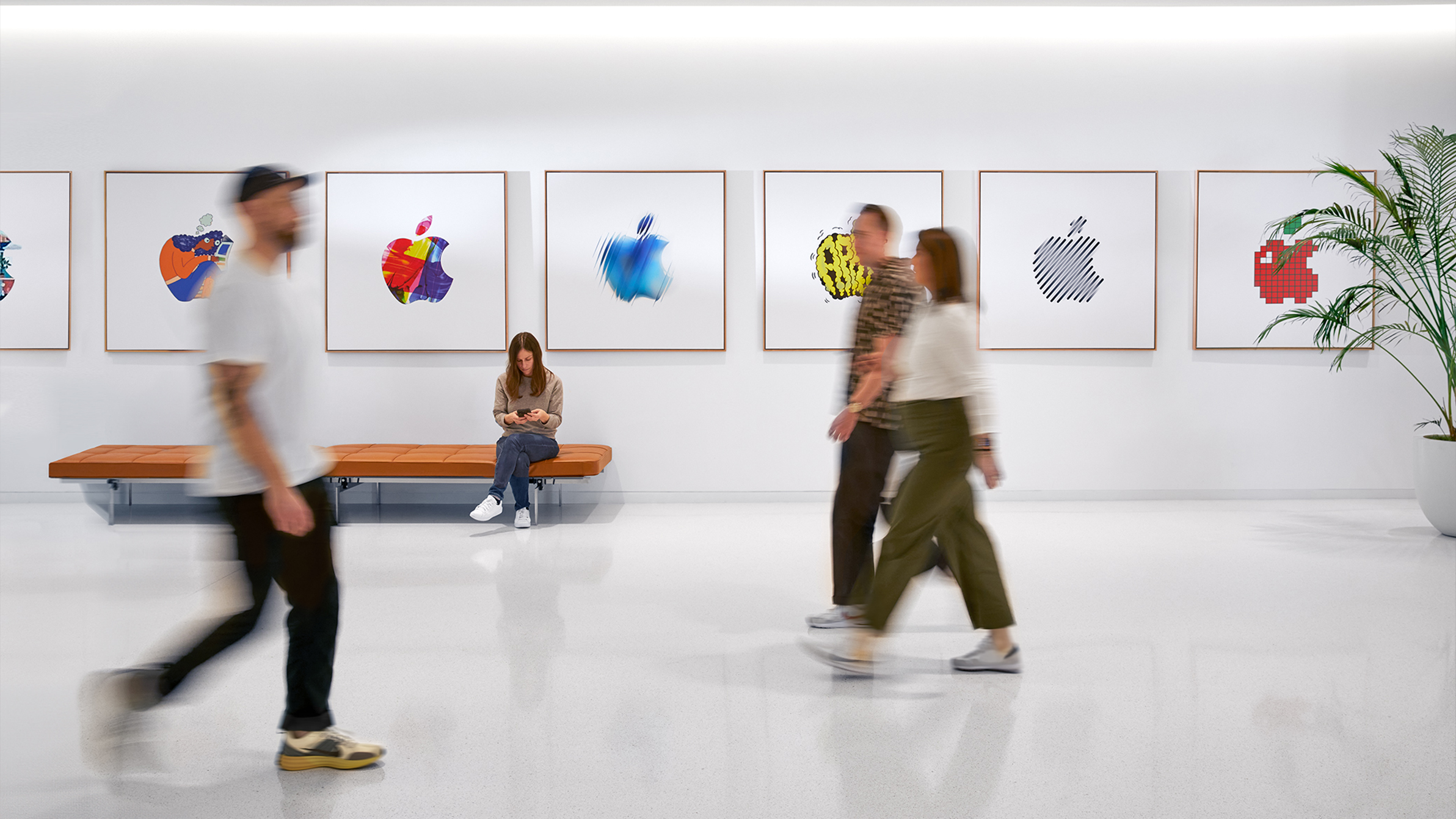 Three Apple colleagues walking past an interior wall with colorful Apple logos, and a fourth employee sitting on a bench.