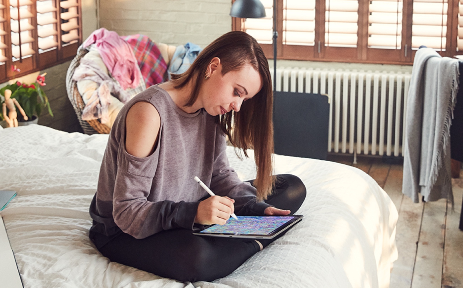 A female Apple Store employee sits on her bed at home while using her Apple Pencil and iPad to draw.