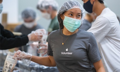 Apple 자원봉사 티셔츠를 입고 있는 Apple 인턴이 자원봉사 이벤트에서 물건을 포장하면서 옆을 바라보며 미소 짓고 있음.