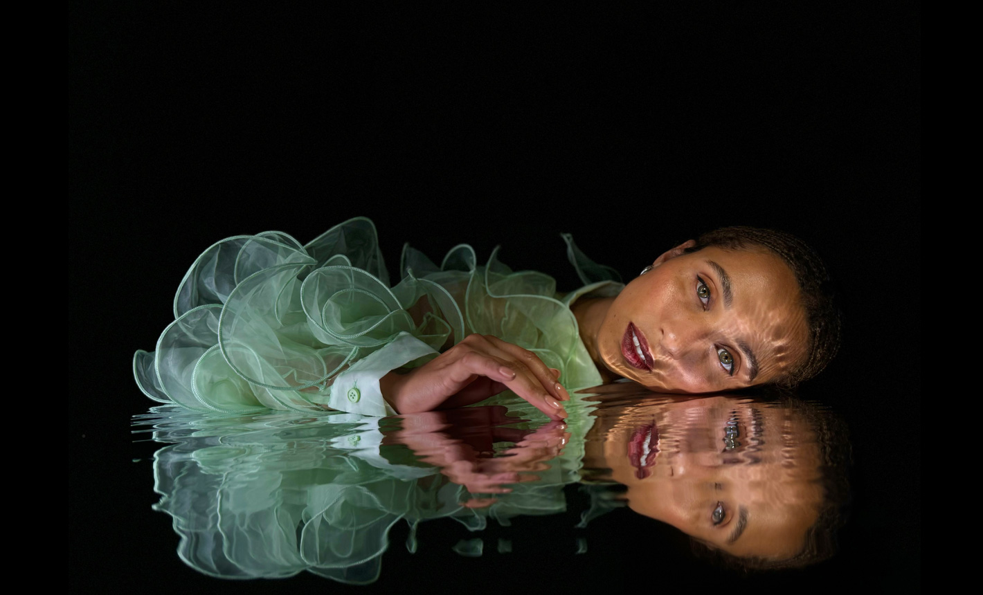 Foto con delicados detalles tomada con un iPhone 15 Pro que muestra a una mujer recostada cerca de su reflejo en aguas onduladas sobre un fondo negro