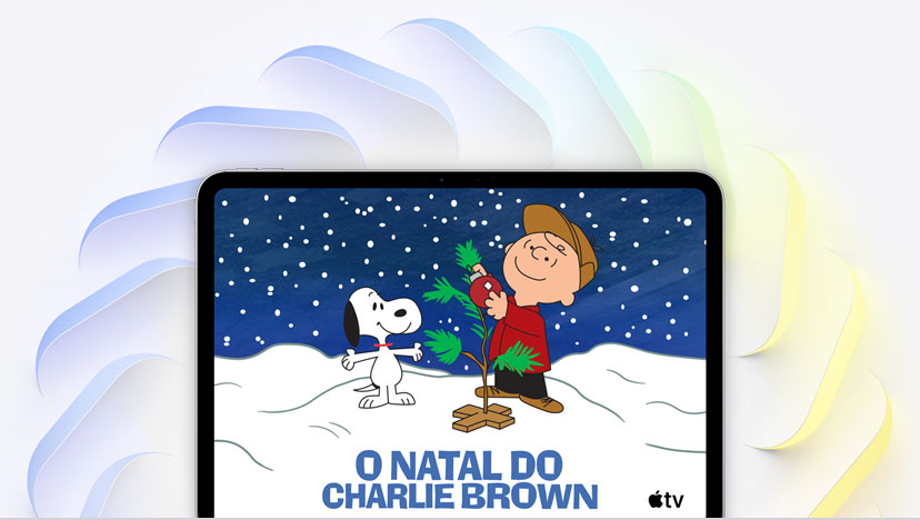 iPad Pro de 11 polegadas prateado, tela com a imagem do Charlie Brown decorando uma pequena árvore de Natal, o Snoopy olhando para a árvore, a neve caindo e o logotipo do Apple TV no canto inferior direito. Diversos quadrados angulares com bordas arredondadas nas cores azul, verde e amarela se desdobram por trás