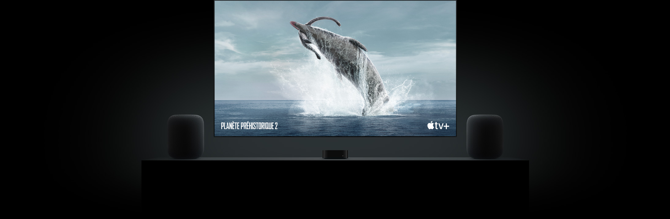 Grand téléviseur à écran plat présentant l’image nette d’un dinosaure de Planète préhistorique. L’appareil est suspendu au-dessus d’une Apple TV et encadré de deux HomePod posés sur un meuble de salon.