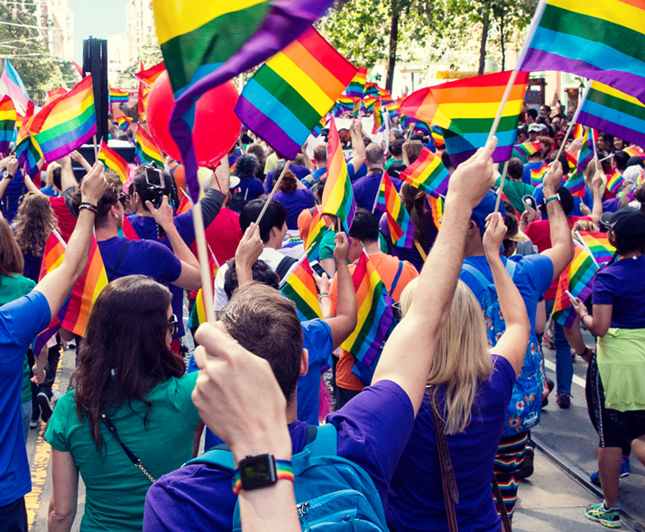 Photo of a crowd of people in a parade waving rainbow flags. 