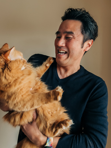 Un ritratto fotografico di Tetsu che sorride mentre osserva e tiene in braccio il suo gatto.