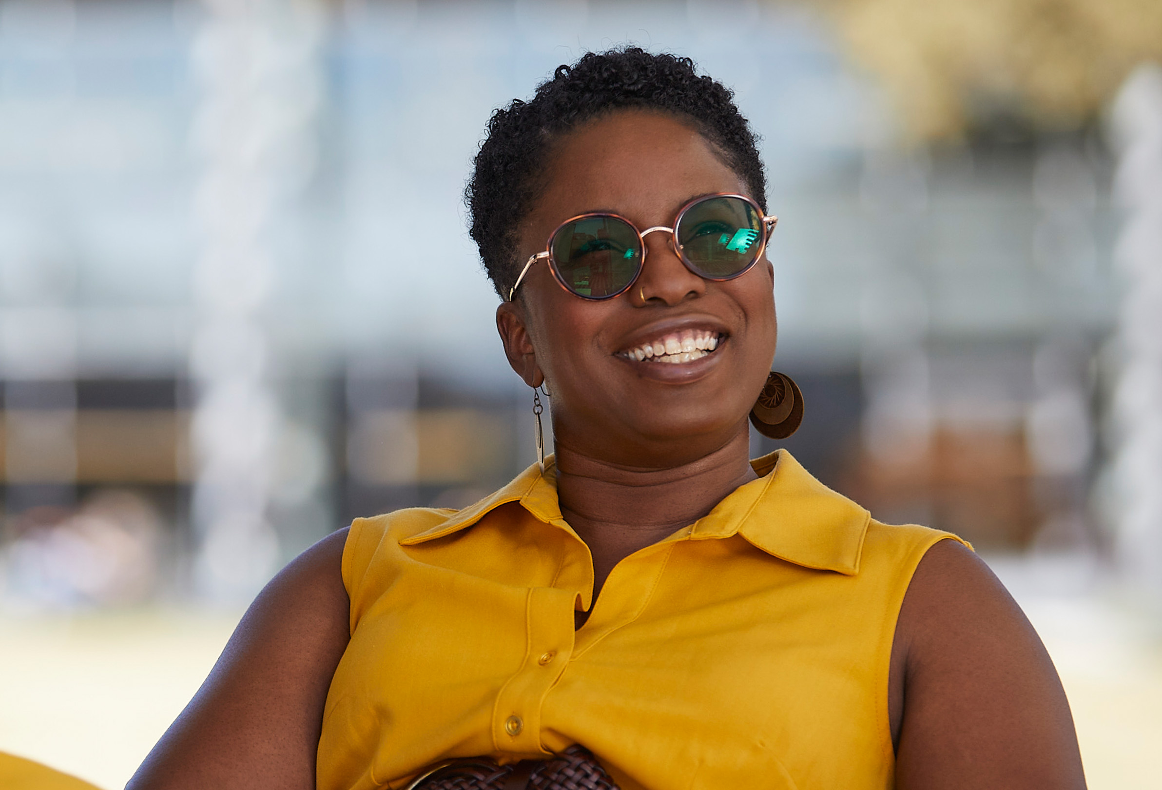 Zaměstnankyně společnosti Apple s nasazenými slunečními brýlemi sedí venku a usmívá se.