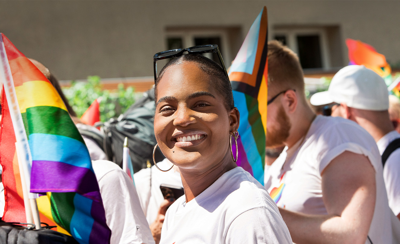 A smiling Apple employee at an Apple Pride event in a crowd of employees holding Pride flags and wearing Pride T-shirts. 