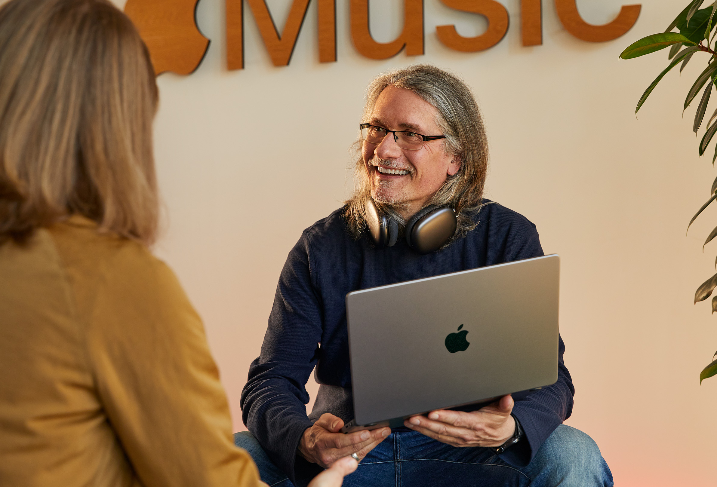 Two Apple employees in conversation, one holding a MacBook and sitting in front of a wall with the Apple Music logo on it.