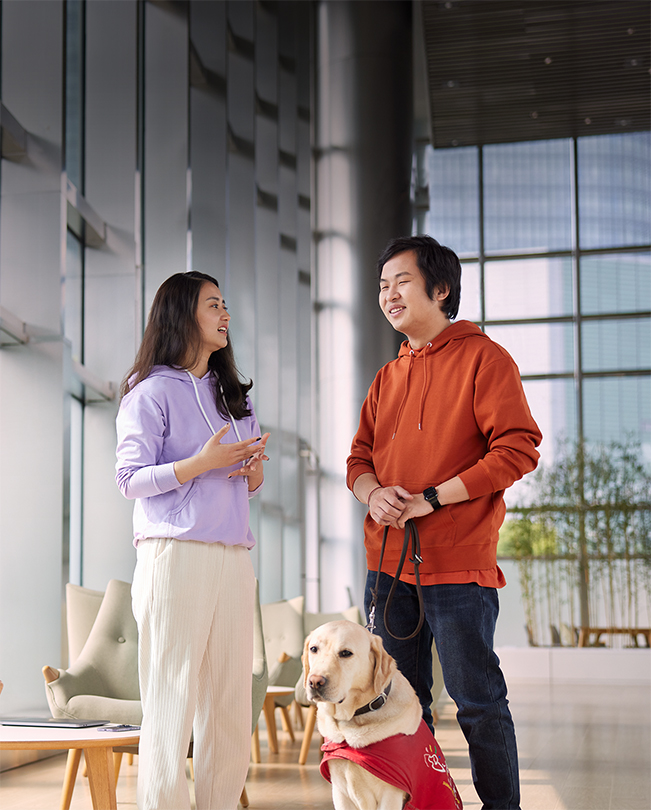 Dos empleados de Apple hablando en una zona común con mucha luz natural; uno de ellos lleva la correa de un perro guía.