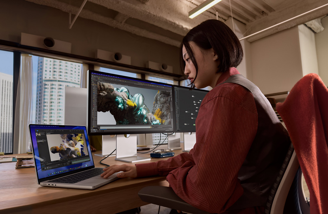 Une personne travaille à un bureau avec un MacBook Pro connecté à un écran externe, les deux écrans affichant un rendu 3D, des bâtiments visibles à travers la fenêtre derrière