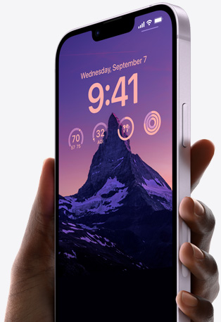 Tangan memegang iPhone 14 berwarna Ungu dengan Layar Terkunci yang dipersonalisasi serta memperlihatkan foto gunung berpencahayaan rendah, waktu, dan widget