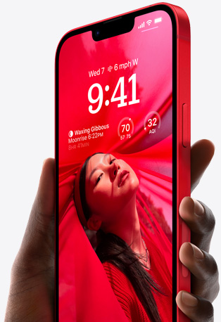 Tangan memegang iPhone 14 berwarna Red dengan Layar Terkunci yang dipersonalisasi serta memperlihatkan foto orang berpakaian serba merah, waktu, dan widget