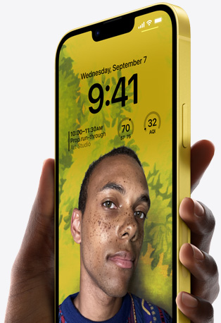 Tangan memegang iPhone 14 berwarna Kuning dengan Layar Terkunci yang dipersonalisasi serta memperlihatkan foto seseorang dengan latar belakang kuning, waktu, dan widget