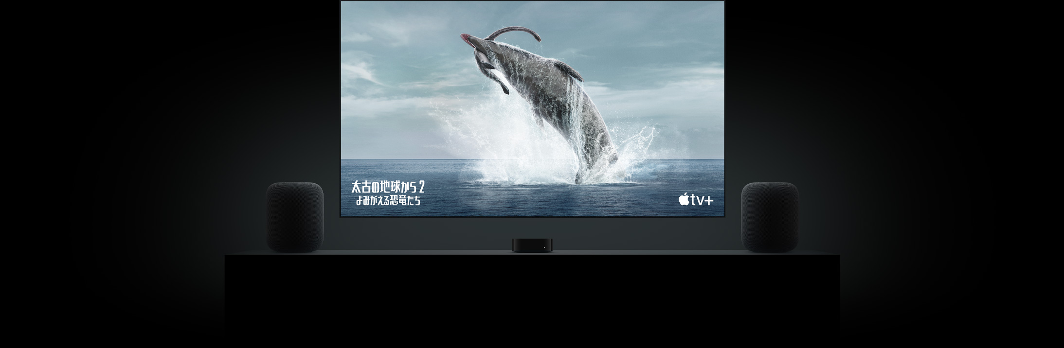 フラットテレビの大きなスクリーン上に「太古の地球から〜よみがえる恐竜たち〜」の恐竜の鮮明な映像が映し出されている。リビングルームのキャビネットの上に置かれた2台のHomePodスピーカー。その間にApple TVが配置され、その上にテレビが掛けられている