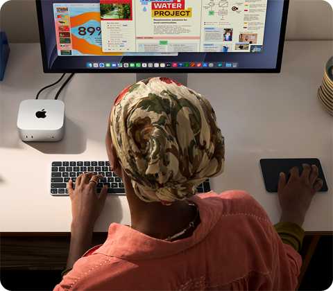 Žmogus rašo klaviatūra prie stalo, ant kurio matomi „Mac mini“, prie jo prijungtas „Studio Display“, „Magic Keyboard“, „Magic Trackpad“ ir įvairūs daiktai.