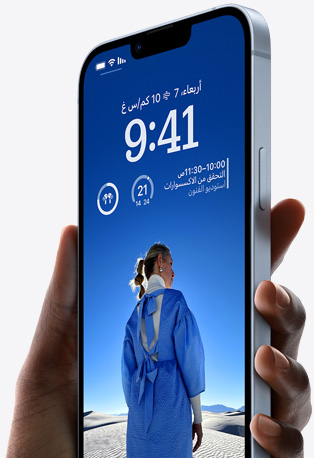 يد تحمل iPhone 14 باللون الأزرق تظهر عليه شاشة قفل مخصصة تضم صورة شخص يرتدي ملابس باللون الأزرق، إلى جانب الوقت، والأدوات.