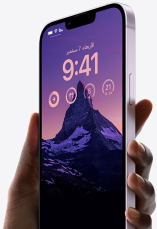 يد تحمل iPhone 14 باللون الليلكي تظهر عليه شاشة قفل مخصصة تضم صورة لجبل بإضاءة خافتة، إلى جانب الوقت، والأدوات