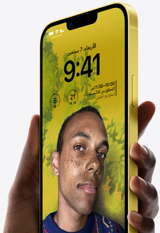 يد تحمل iPhone 14 باللون الأصفر تظهر عليه شاشة قفل مخصصة تضم صورة لشخص مع خلفية صفراء، إلى جانب الوقت، والأدوات