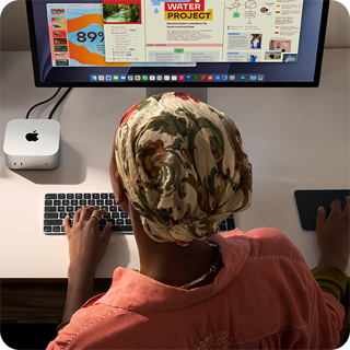 شخص يطبع وهو جالس إلى مكتب مع جهاز Mac mini، شاشة Studio Display متصلة، لوحة مفاتيح ماجيك، لوحة تعقب ماجيك، أغراض متنوعة