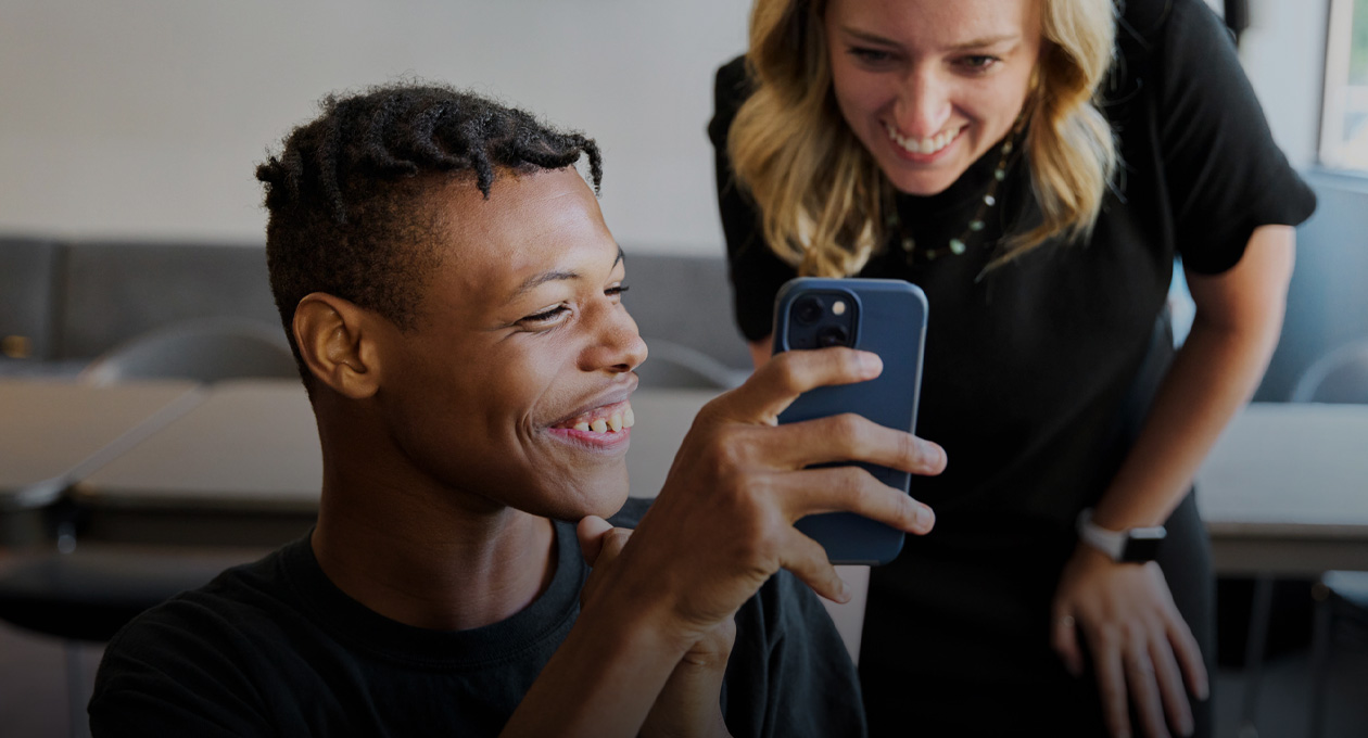 Foto di un giovane ipovedente che sorride e mostra il display del suo iPhone a una donna sorridente in piedi dietro la sua spalla sinistra