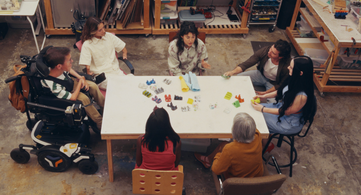 Un gruppo di persone con diverse esigenze di mobilità sedute insieme a un tavolo mentre guardano una serie di accessori colorati e adattabili per smartphone