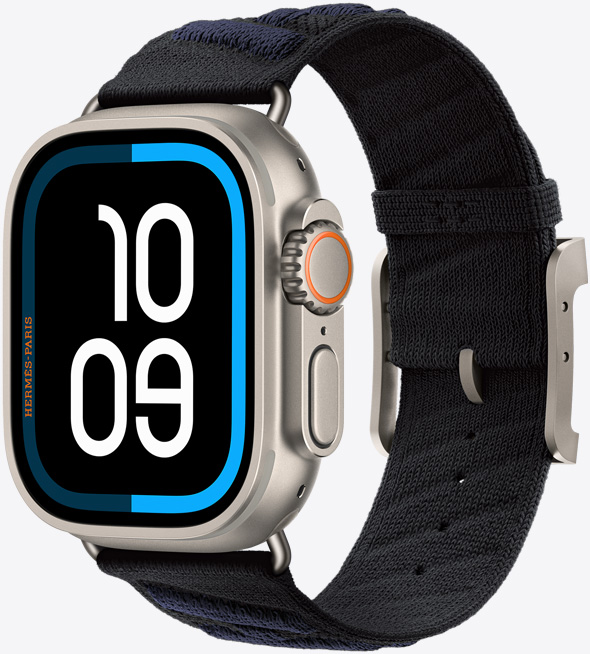 Apple Watch Hermès Ultra 3, titanium case, natural colour, edge-to-edge display, rounded corners, right side exterior, Digital Crown, microphone, raised side button, Maritime digital watch face (10 09 digits), paired with En Mer band, noir/bleu nuit colour (black with dark blue diagonal textured stripes), knit polyamide yarn material, adjustment holes centred, natural colour titanium buckle and hardware