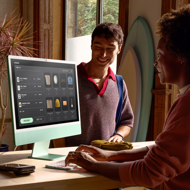 Un employé assis derrière le comptoir d’un magasin commercialisant des planches de surf et d’autres articles utilise un iMac pour finaliser une transaction avec un client se trouvant de l’autre côté du comptoir.