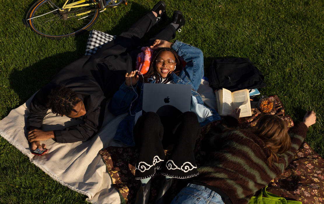 Tres estudiantes universitarios acostados sobre una manta en el parque. Un estudiante tiene un iPhone. Una estudiante tiene un MacBook Air y audífonos Apple. Una estudiante tiene un libro de bolsillo.