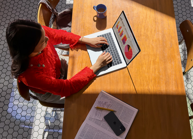 테이블에서 Macbook을 사용하는 사람의 모습. 옆에 머그잔, 스마트폰, 그리고 서류가 있습니다.