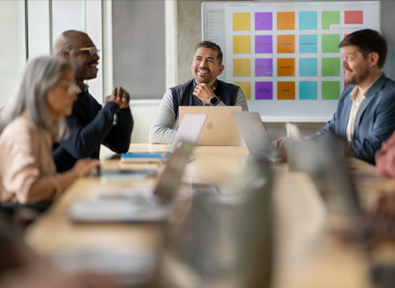Several university staff talk around a table, each with a MacBook open, and a project board on the wall