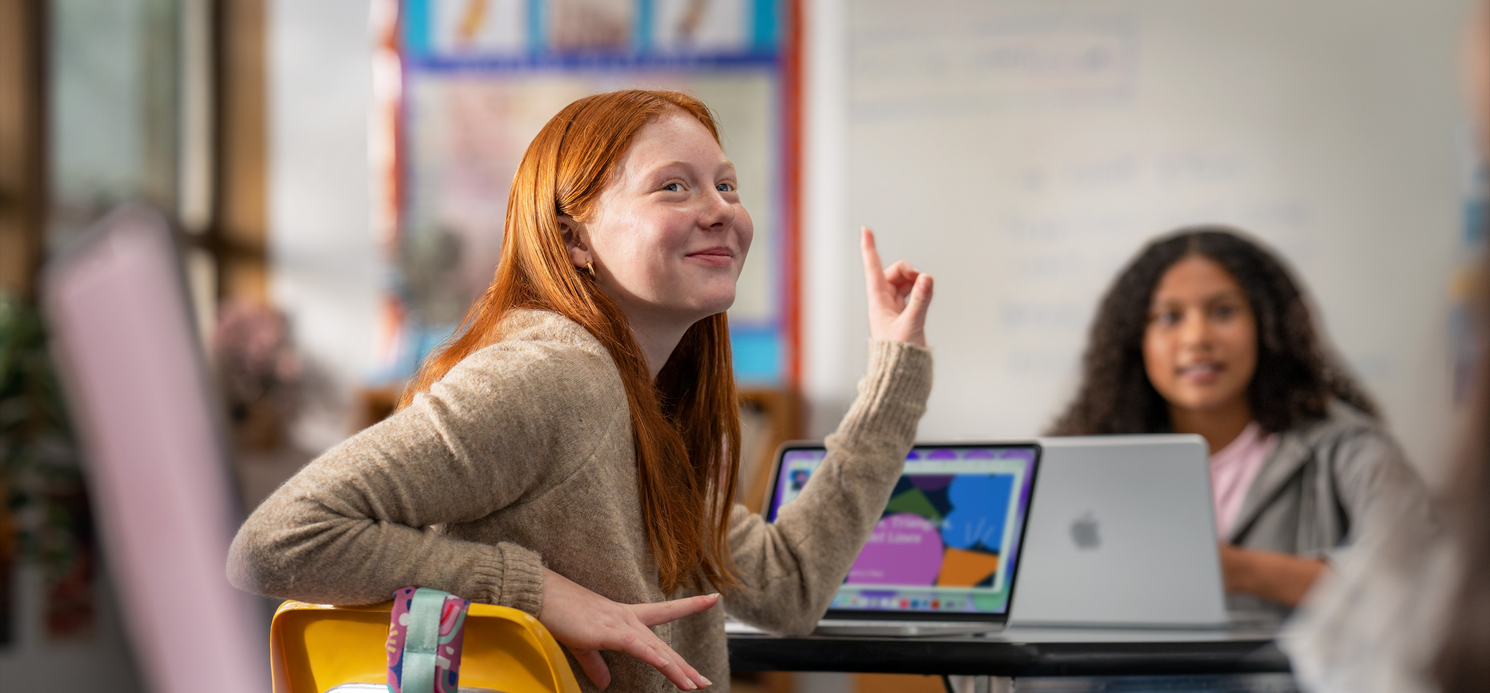 Une élève assise devant un MacBook dans une salle de classe lève la main pour répondre à une question, suggérant la facilité d’utilisation de l’appareil et la participation active de l’élève