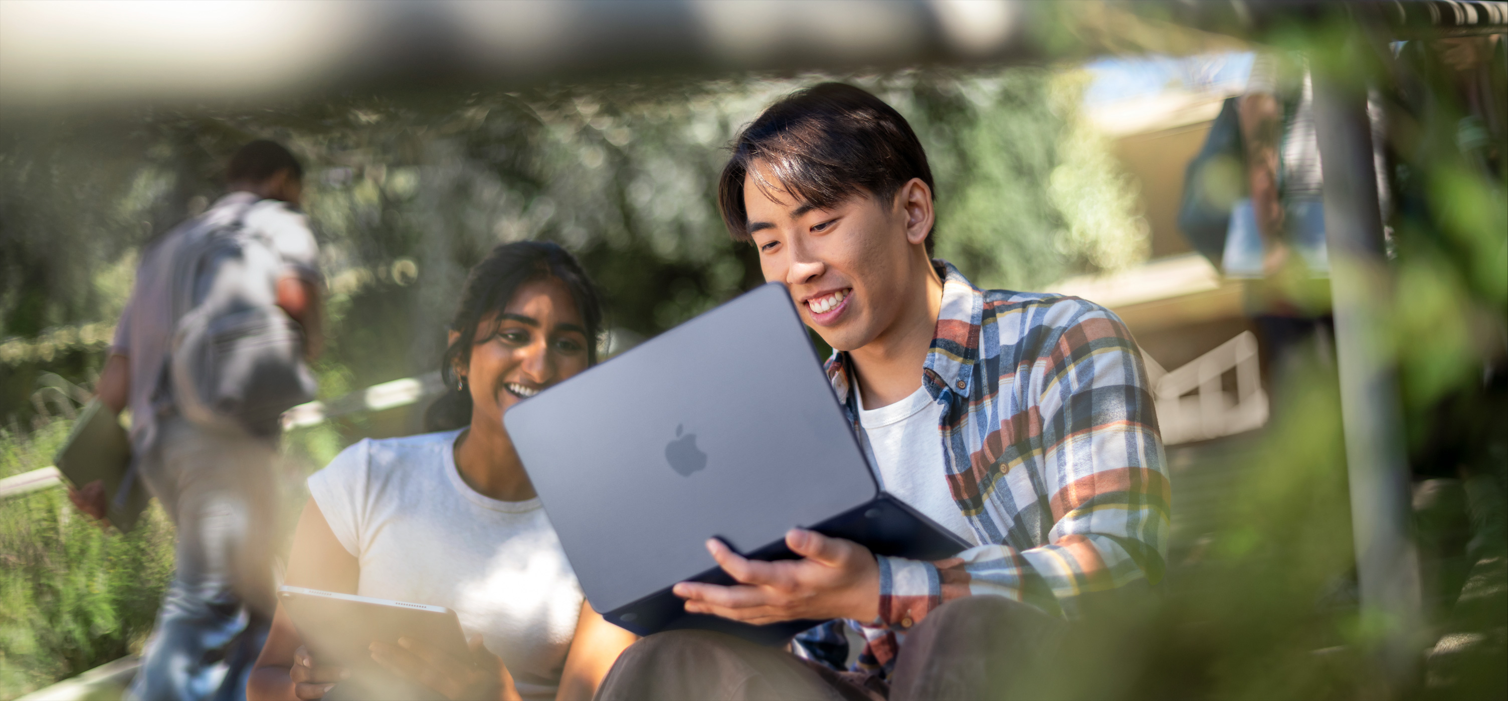 Un étudiant et une étudiante souriants travaillent ensemble sur un MacBook en extérieur, avec l’appareil posé sur les genoux de l’étudiant