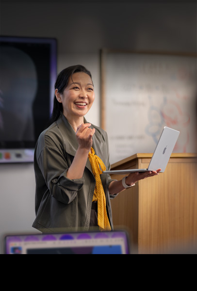Une professeure donne un cours dans un amphithéâtre tout en tenant un MacBook argent, illustrant les performances et la grande autonomie de l’appareil