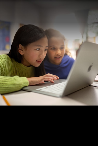 Deux jeunes élèves assises à un bureau travaillent ensemble en classe, les yeux rivés sur un MacBook