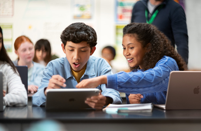 Two students point to an iPad screen in excitement, while a MacBook is also open on the desk