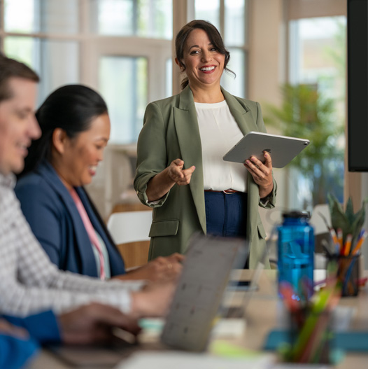 An educator holds an iPad while presenting to two colleagues who are working on Mac in an office setting