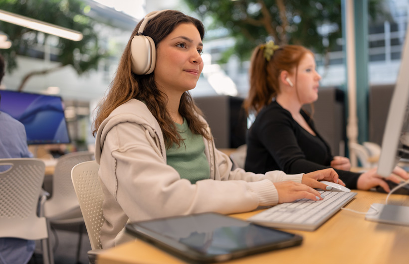 Une étudiante portant des AirPods Max est assise devant un ordinateur et tape sur le clavier avec sa tablette à portée de main, alors qu’une autre étudiante travaille à ses côtés.