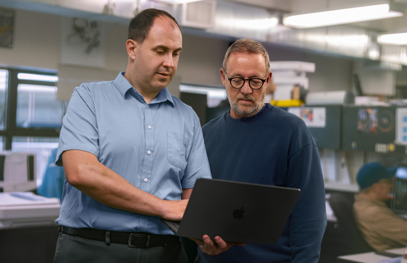 Deux hommes à un poste de travail regardent un MacBook ensemble, l’un des deux le tenant et l’autre pointant l’écran.