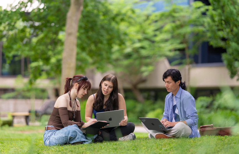 Sur un campus, trois personnes sont assises dans l’herbe, l’une d’elles utilise un MacBook tandis qu’une autre l’observe.