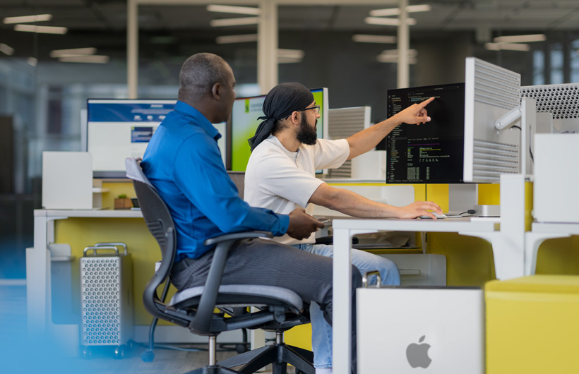 Deux personnes sont assises devant plusieurs moniteurs d’iMac, et l’une d’elles pointe un écran.