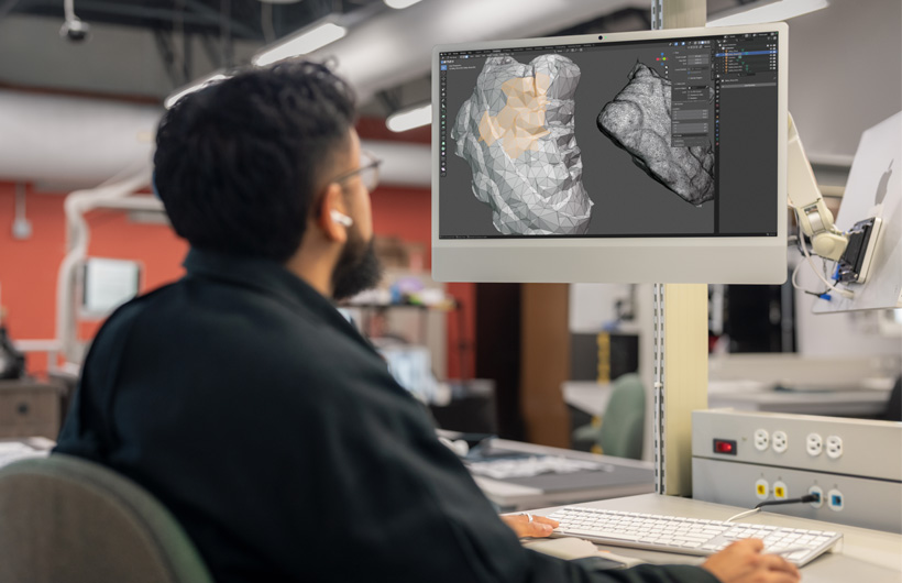 Un étudiant est assis à un bureau et examine deux modèles 3D numérisés affichés côte à côte sur des moniteurs d’iMac.