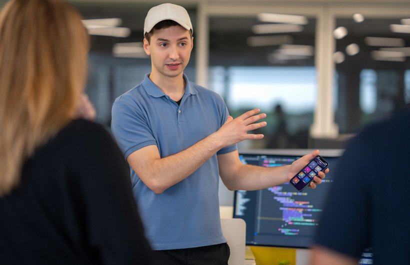 Un étudiant parle à deux personnes et présente son iPhone dont l’écran d’accueil affiche plusieurs apps. Derrière lui, un moniteur affiche un éditeur de code avec du texte coloré surligné pour montrer la syntaxe.
