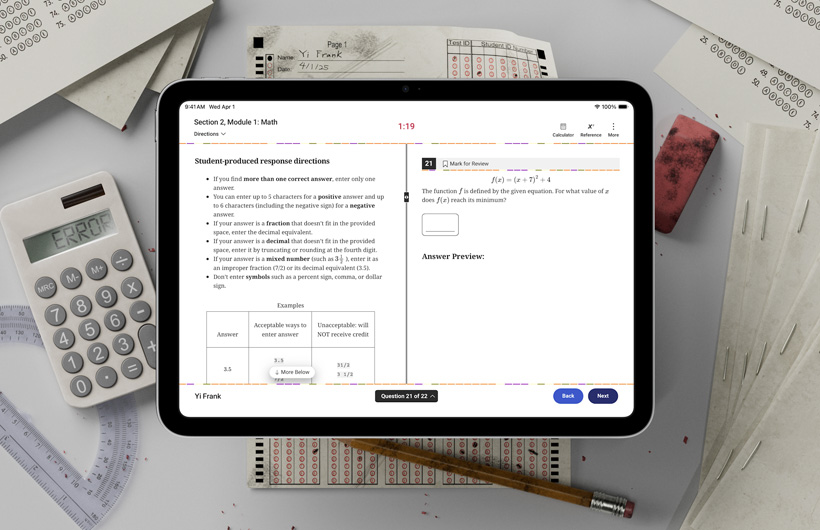 An iPad sits on a desk among physical testing forms and school supplies. On screen, there are instructions for completing the math portion of a standardised test.