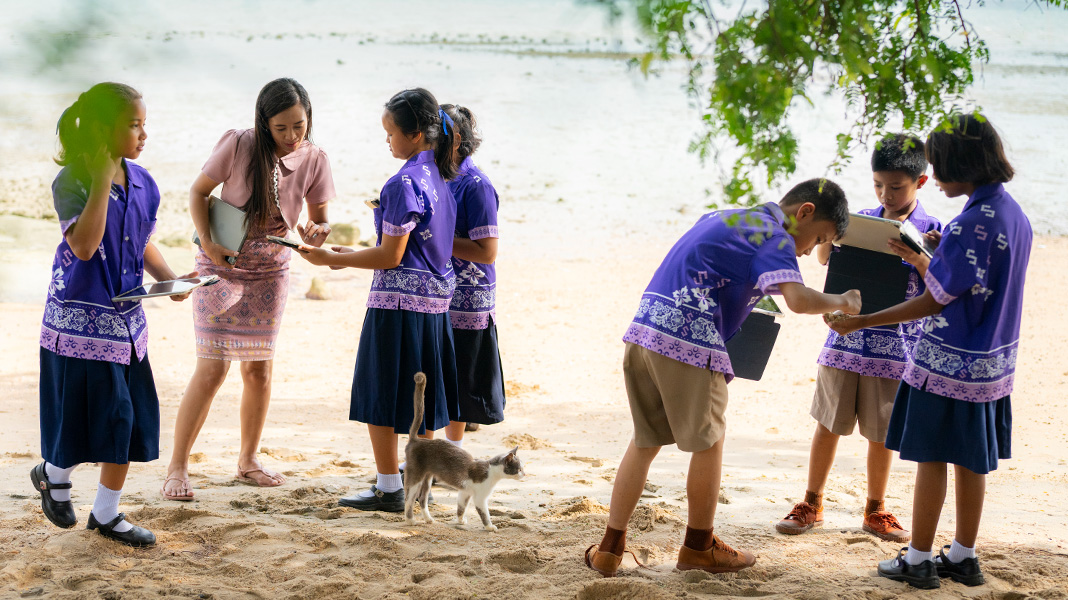 Seis alunos do ensino fundamental, com uniformes roxos coloridos, estão na areia da praia segurando aparelhos iPad. Uma professora ajuda uma aluna com um iPad, e um gato caminha entre elas
