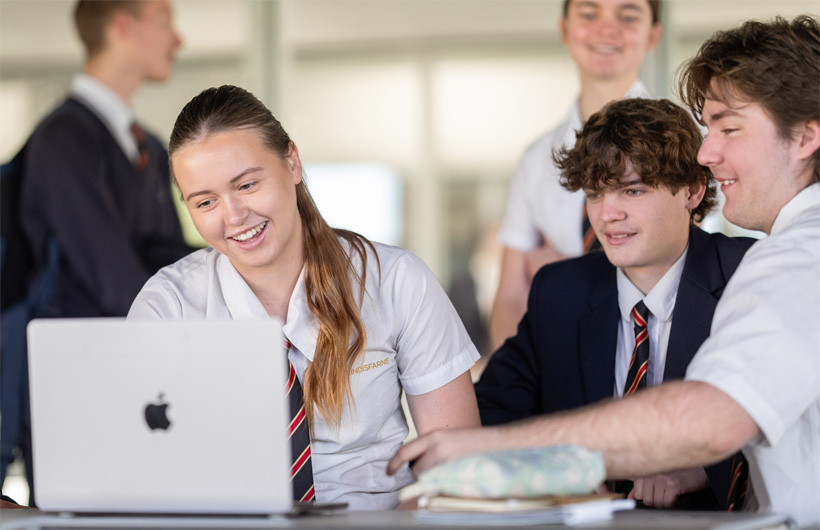 A group of students in school uniforms gather around a MacBook, smiling and collaborating.