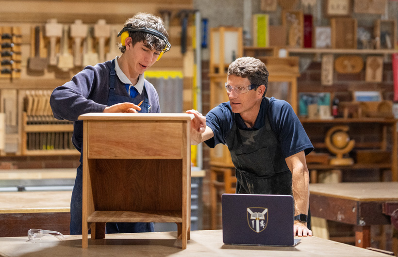 A woodworking instructor and student examine a small wooden cabinet. A branded school laptop sits on the workbench in front of them.