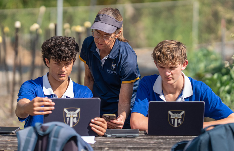 A teacher holds an iPhone and two students review information on their iPad while sitting on a bench in a garden.