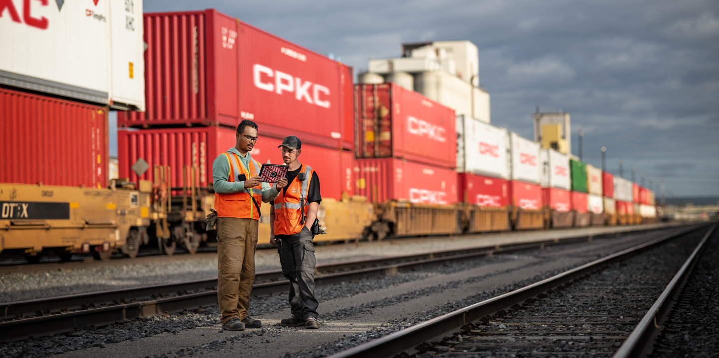 2人の鉄道員が、操車場で停車中の鉄道コンテナの横に立ってiPadを見ている