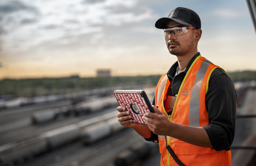 Ein Bahnmitarbeiter, umgeben von großen Waggons, steht mit einem iPad in der Hand und schaut nach vorne