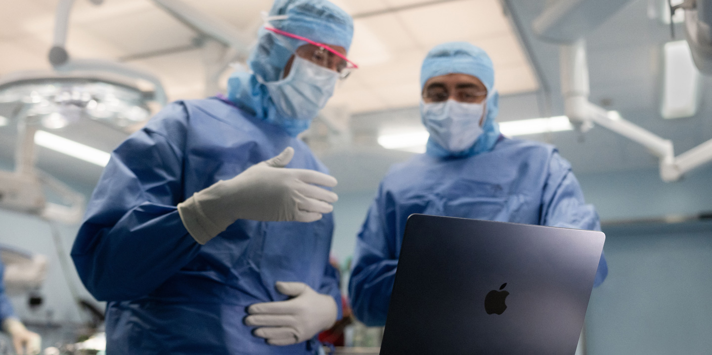Two surgeons in the operating room look at a MacBook, and one gestures with their hand while the other observes